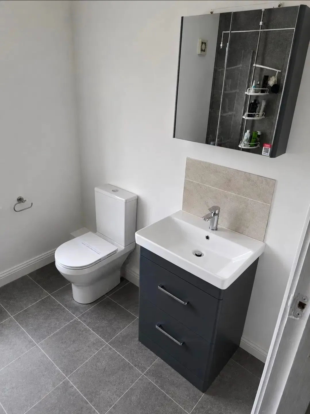 Completed bathroom installation with a white toilet, dark grey vanity unit with white basin and chrome tap, grey tiled floor and a mirrored cabinet above